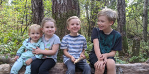 Four grinning children sitting on a log surrounded by old-growth forest.